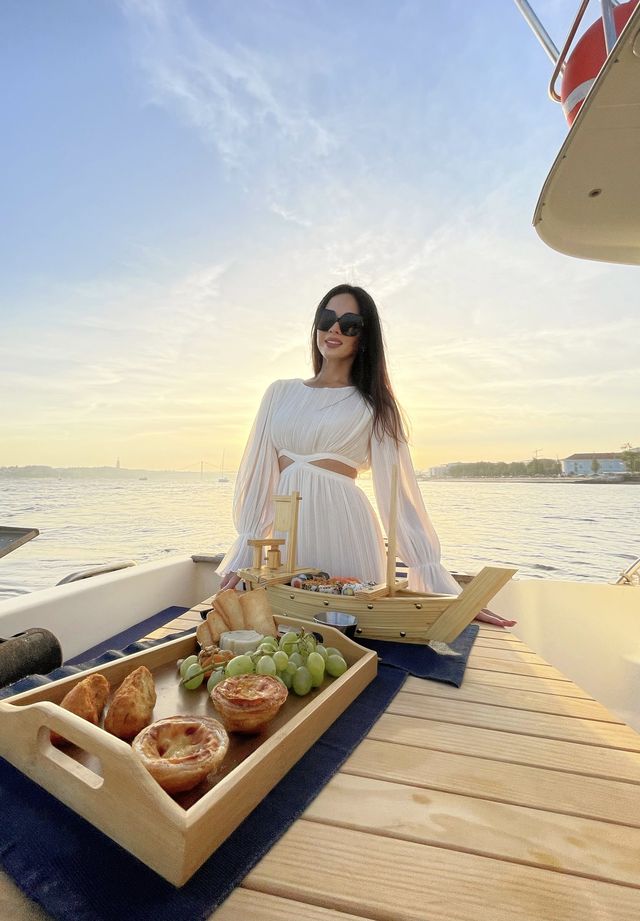 Karolina in a white outfit on a boat deck at golden hour with a picnic tray of bread and fruit