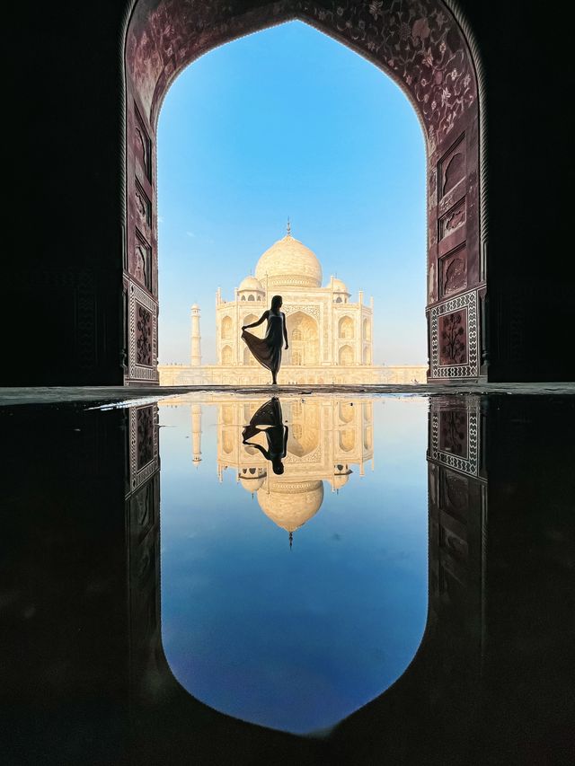 The Taj Mahal and its reflection framed by a dark archway, with a silhouetted figure in a flowing dress